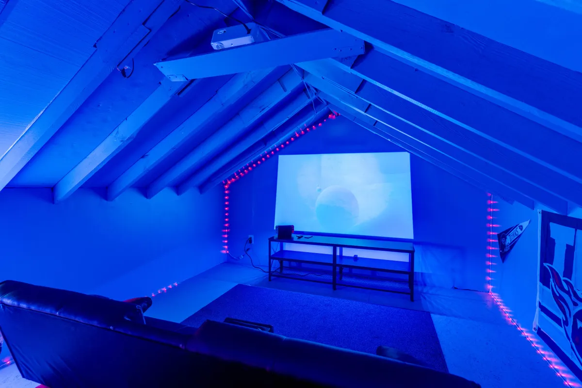 A modern home theatre setup with a large projector screen dominating the wall, LED accent lights setting the mood, and a comfy sofa for cozy movie nights. The space exudes the inviting ambiance of a private cinema.