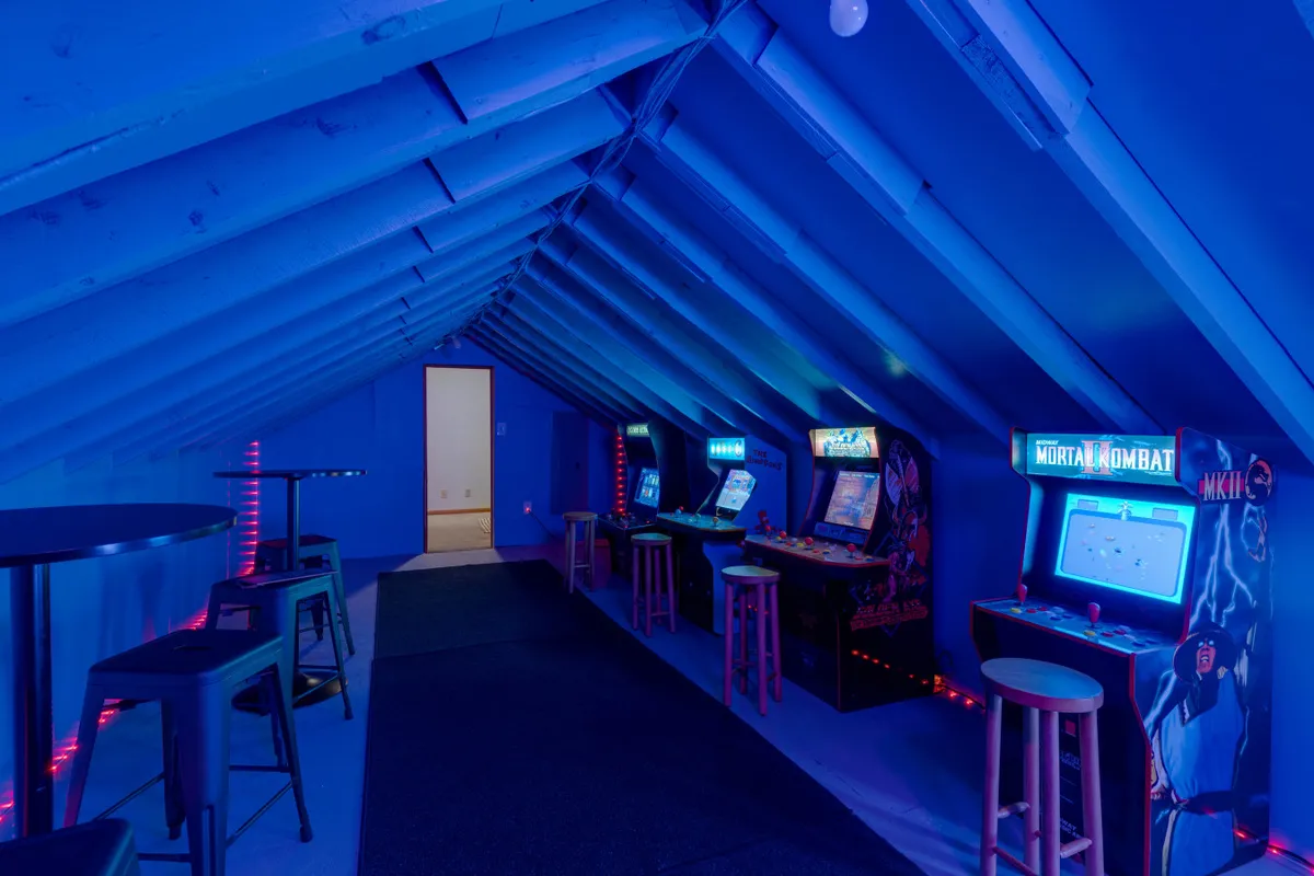 A vibrant game room filled with arcade machines lined against a wall, atmospheric lighting, and bar tables for socializing. The lighting effects and the presence of multiple game stations create a lively, fun-filled vibe perfect for competitive gaming and group entertainment.