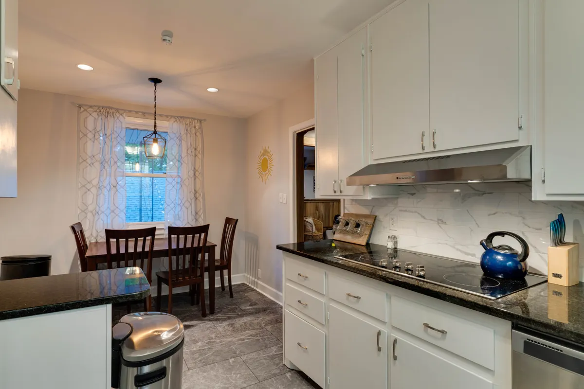 A sunlit kitchen and dining area featuring a kitchen counter, dining table, and a window adorned with curtains. The sun decoration on the wall and ample natural light create a warm, welcoming vibe, ideal for family meals or casual gatherings.