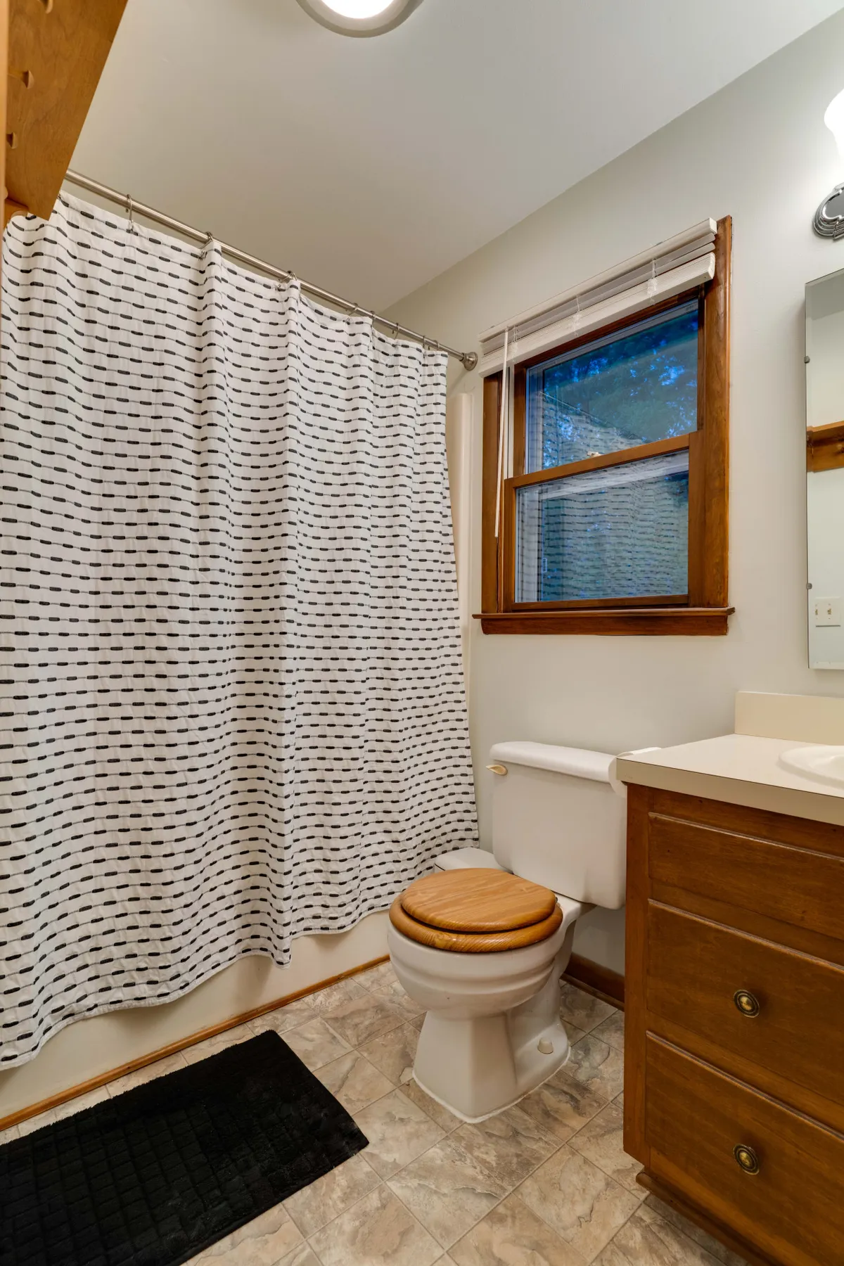 A bathroom scene with a shower curtain, toilet, vanity with sink, and a window. The space is tidy, calm, and welcoming, suggesting cleanliness and comfort.