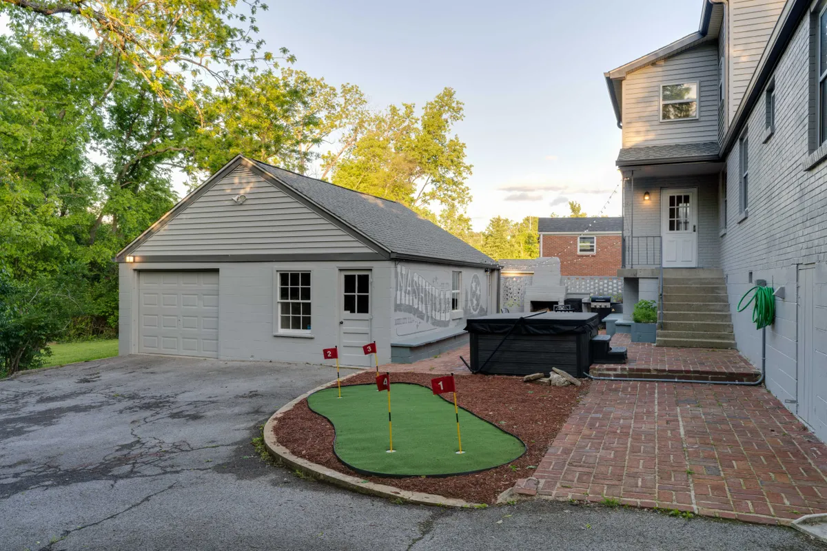This image captures the backyard patio area, complete with a golf putting green, garage, and staircase leading up to the house. The space is set up for outdoor gatherings with a playful, relaxed atmosphere, and ample room for group games or social events.