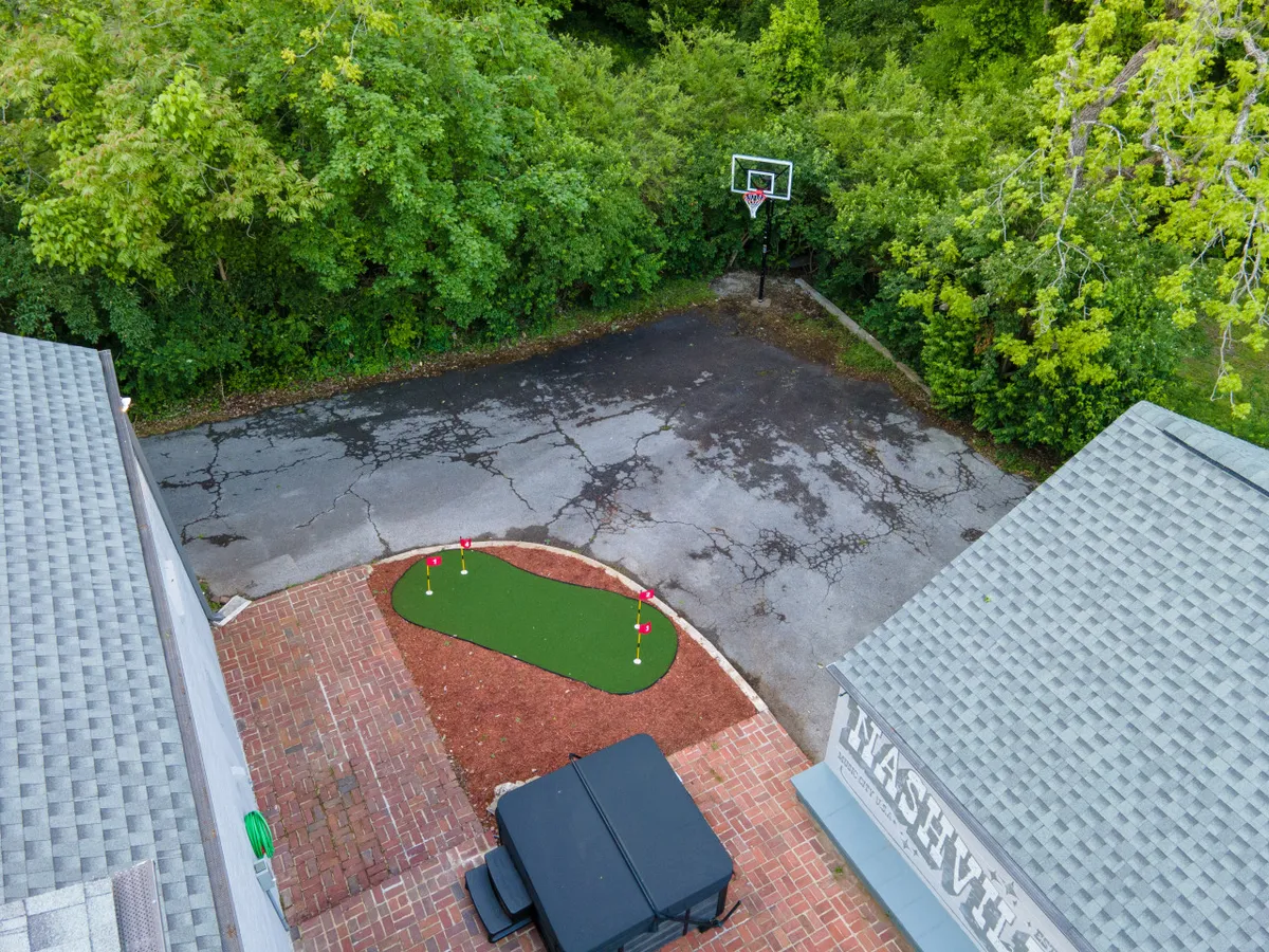 An exterior shot featuring a green putting area for mini-golf, a basketball hoop nearby, a brick pathway, and surrounding trees. The cheerful outdoor setup signals fun and friendly competition in a relaxed backyard environment.