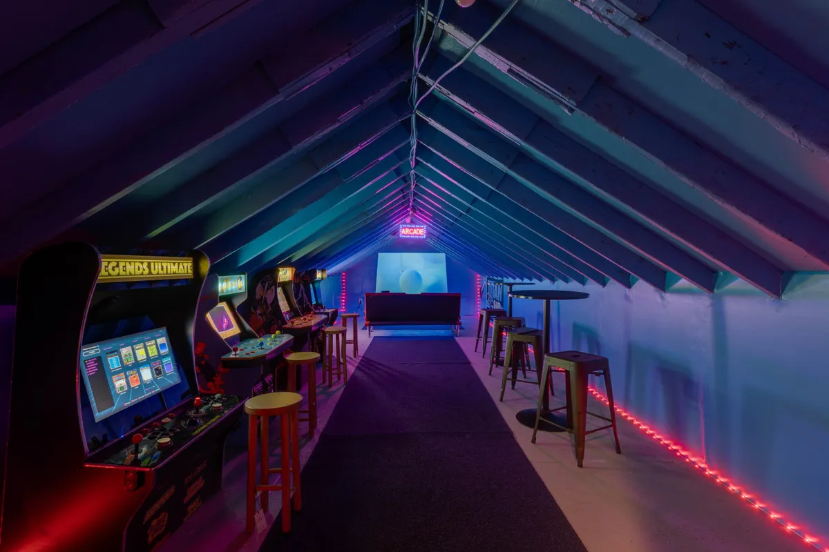 The image captures a vibrant game room filled with arcade machines, neon lights, cozy bar stools, a plush couch, and a cheerful welcome sign, all set up for energetic gaming and socializing.