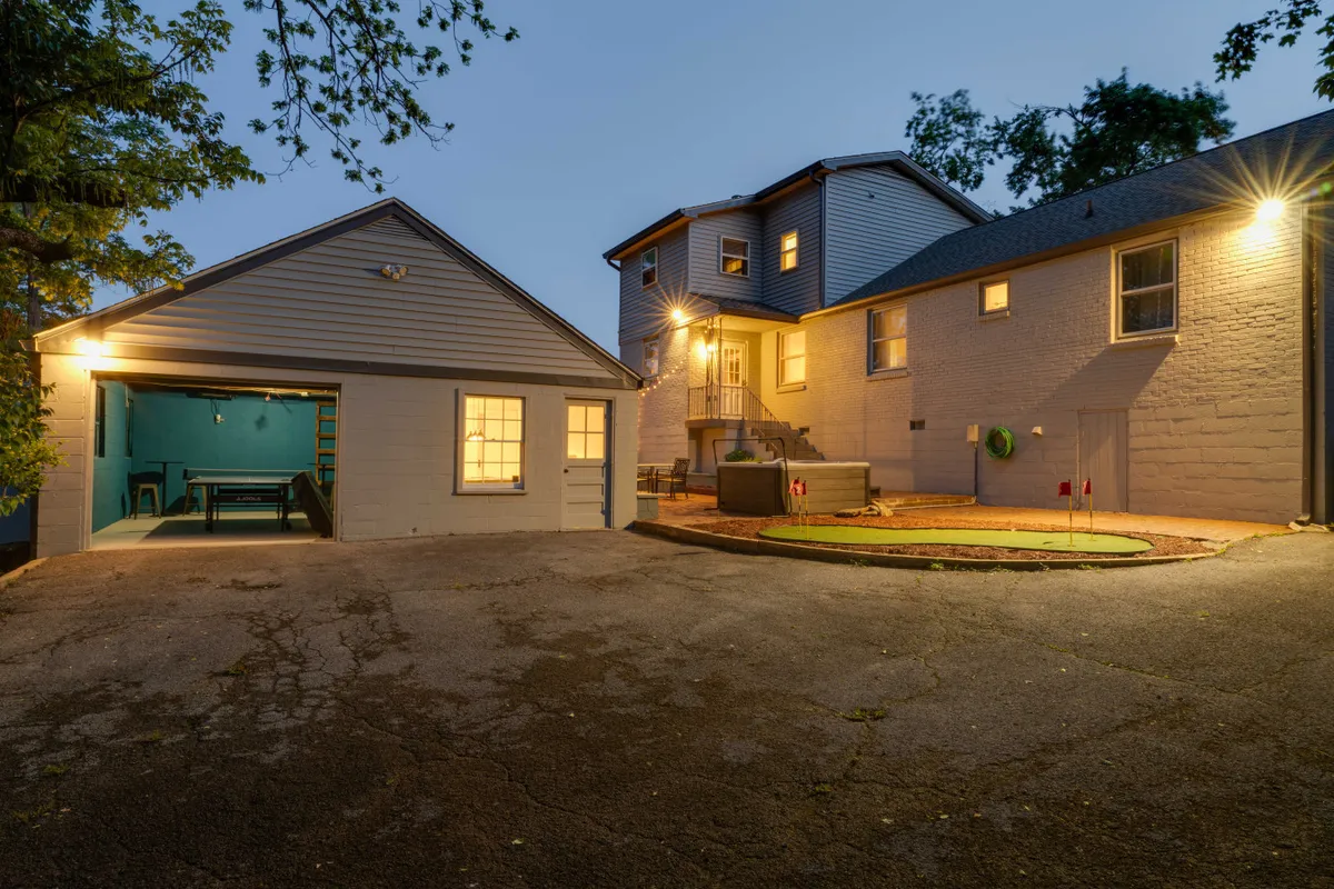 This exterior image showcases the garage entrance, illuminated windows, backyard seating area, and a hot tub. The cozy ambiance suggests an inviting space for evening relaxation and social gatherings outdoors.