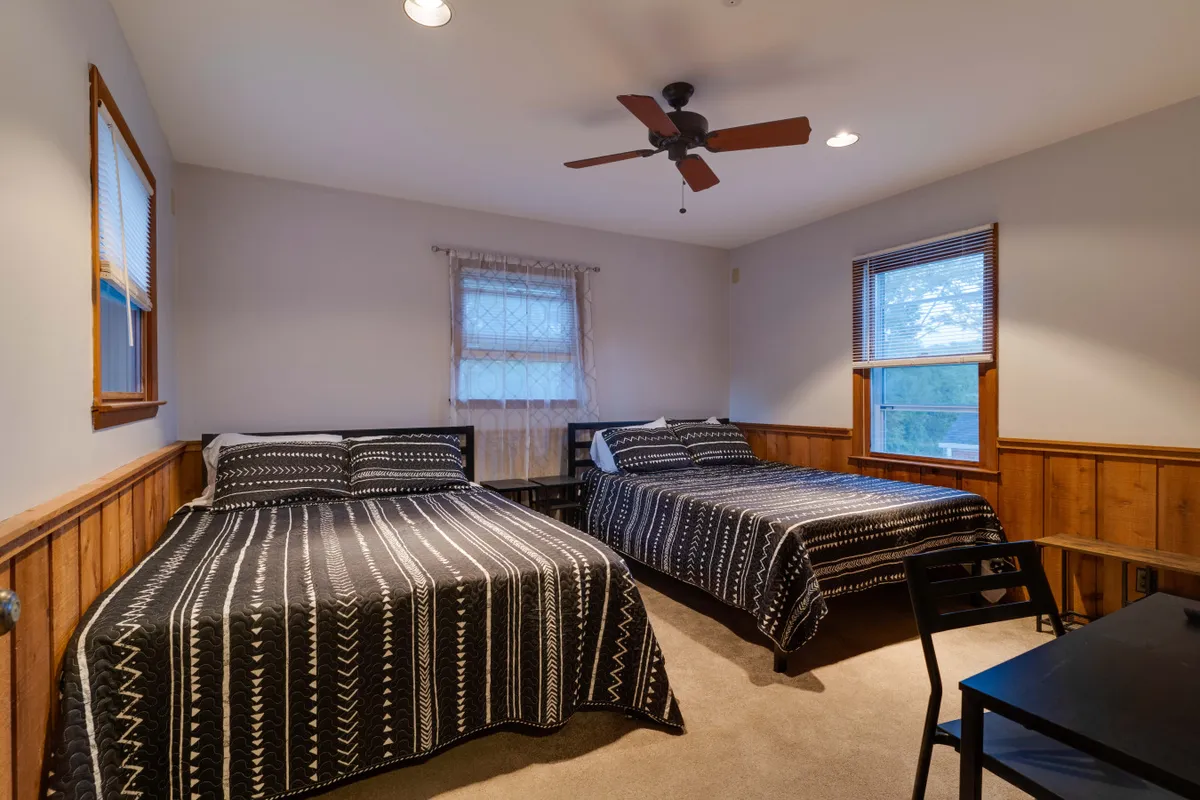 This image features two inviting beds with patterned bedspreads, wooden paneling, a ceiling fan, and windows letting in natural light, creating a tranquil, comfortable atmosphere ideal for rest.
