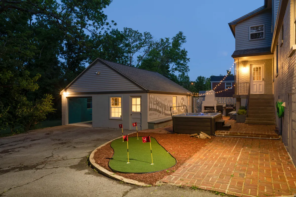 A lively exterior shot showing a putting green, hot tub, outdoor lighting, and garage. It conveys a sense of casual, welcoming space for outdoor games, relaxation, and evening gatherings.