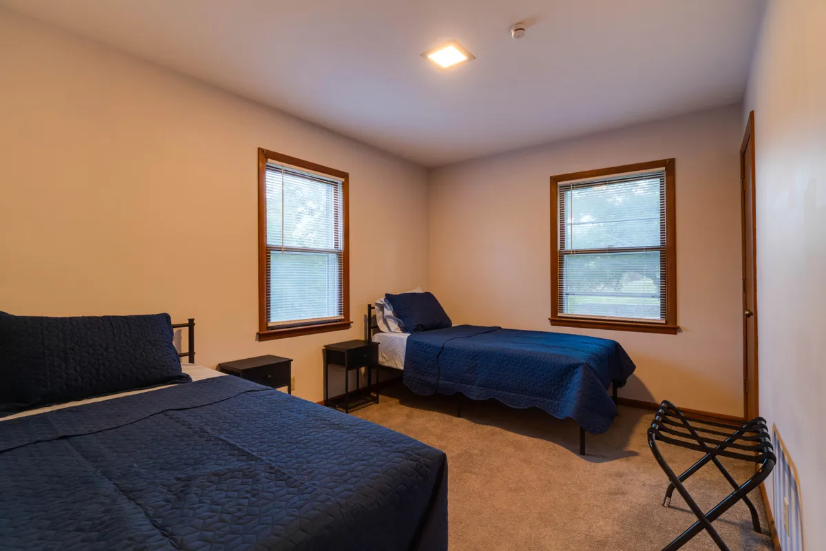 A bright bedroom outfitted with two beds, windows, nightstands, and a folding rack. The room looks comfortable, with soft lighting and a layout that suggests both relaxation and socializing.