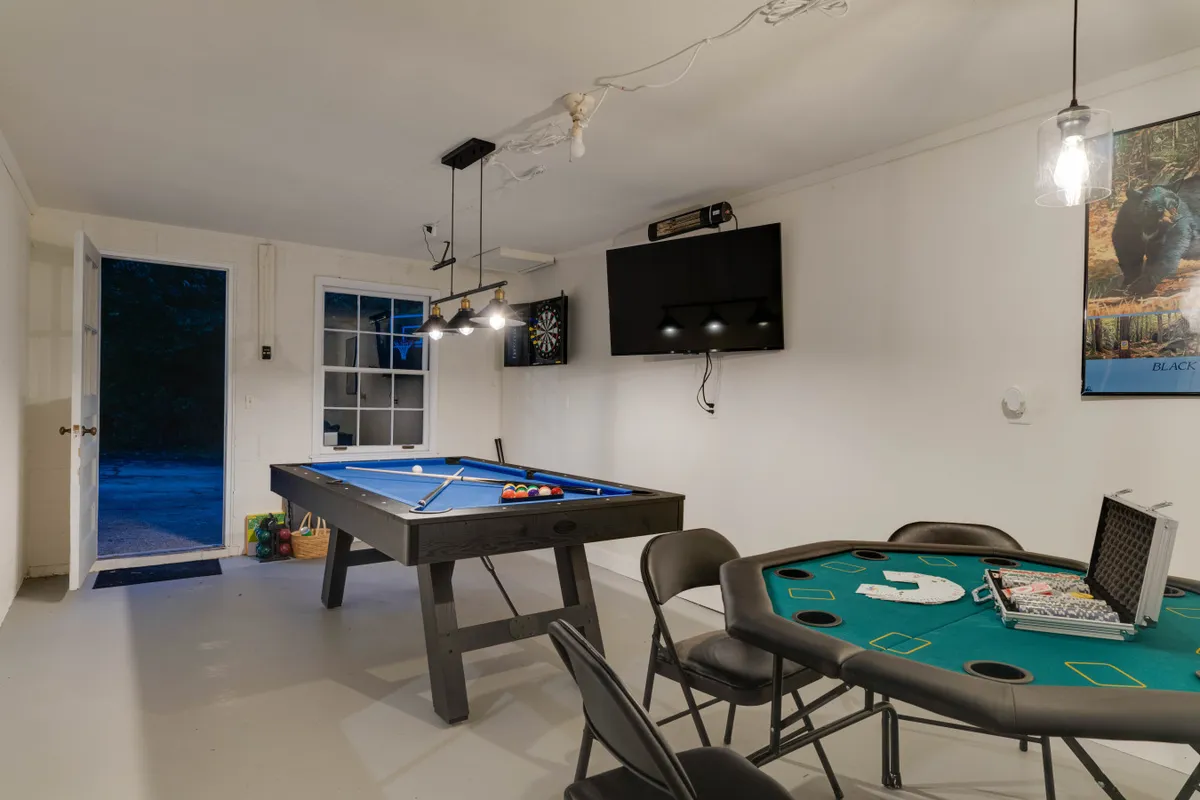 A spacious game room featuring a pool table, a poker/card table, TV, dartboard, and an open door inviting further exploration. The room buzzes with activity, offering multiple gaming options for all ages.