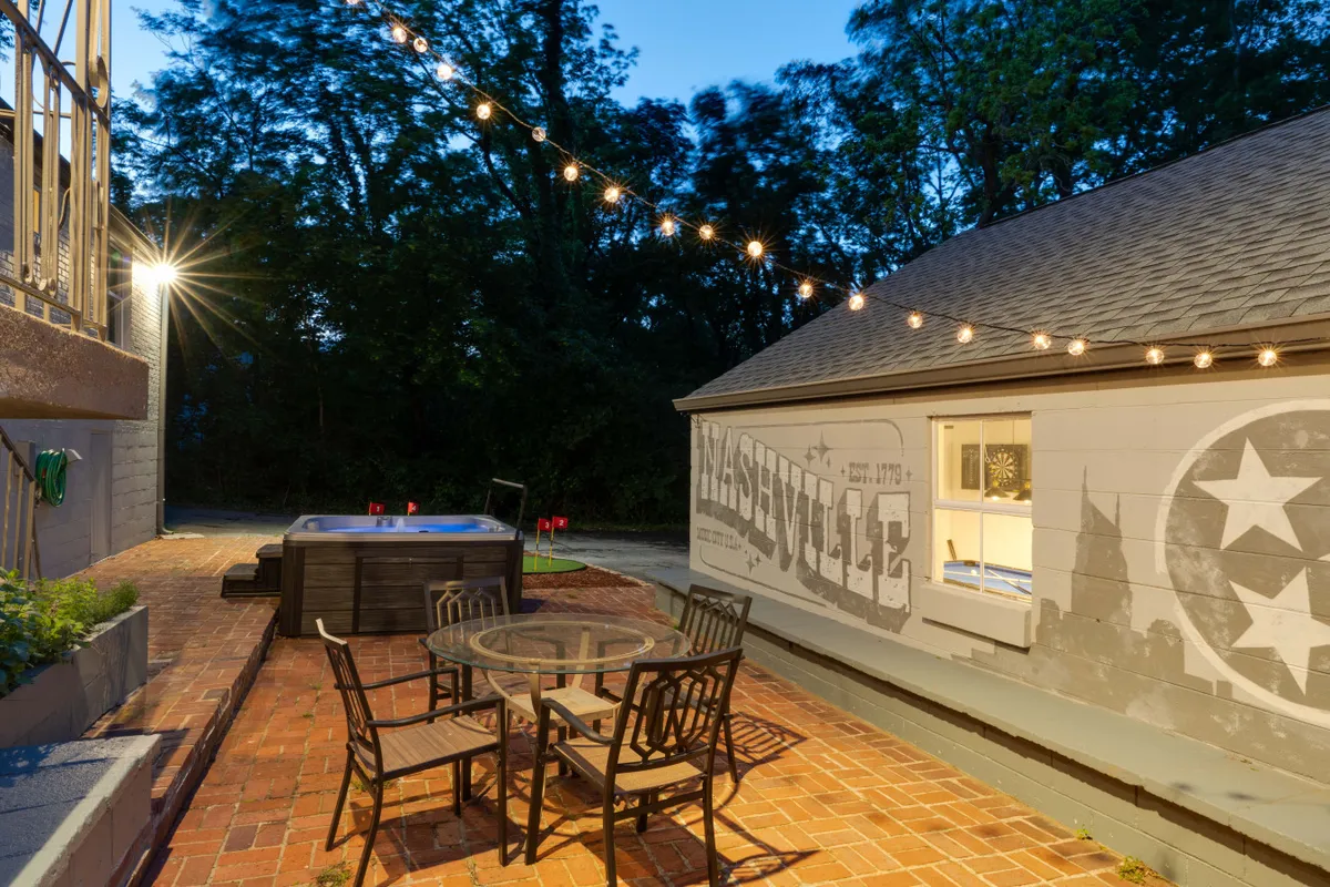 This image presents the deck at dusk, featuring a bubbling hot tub, a festive string of lights overhead, a welcoming outdoor dining set, and a vibrant Nashville mural as a colorful backdrop. Several guests are enjoying the space, highlighting areas for relaxation and celebration.